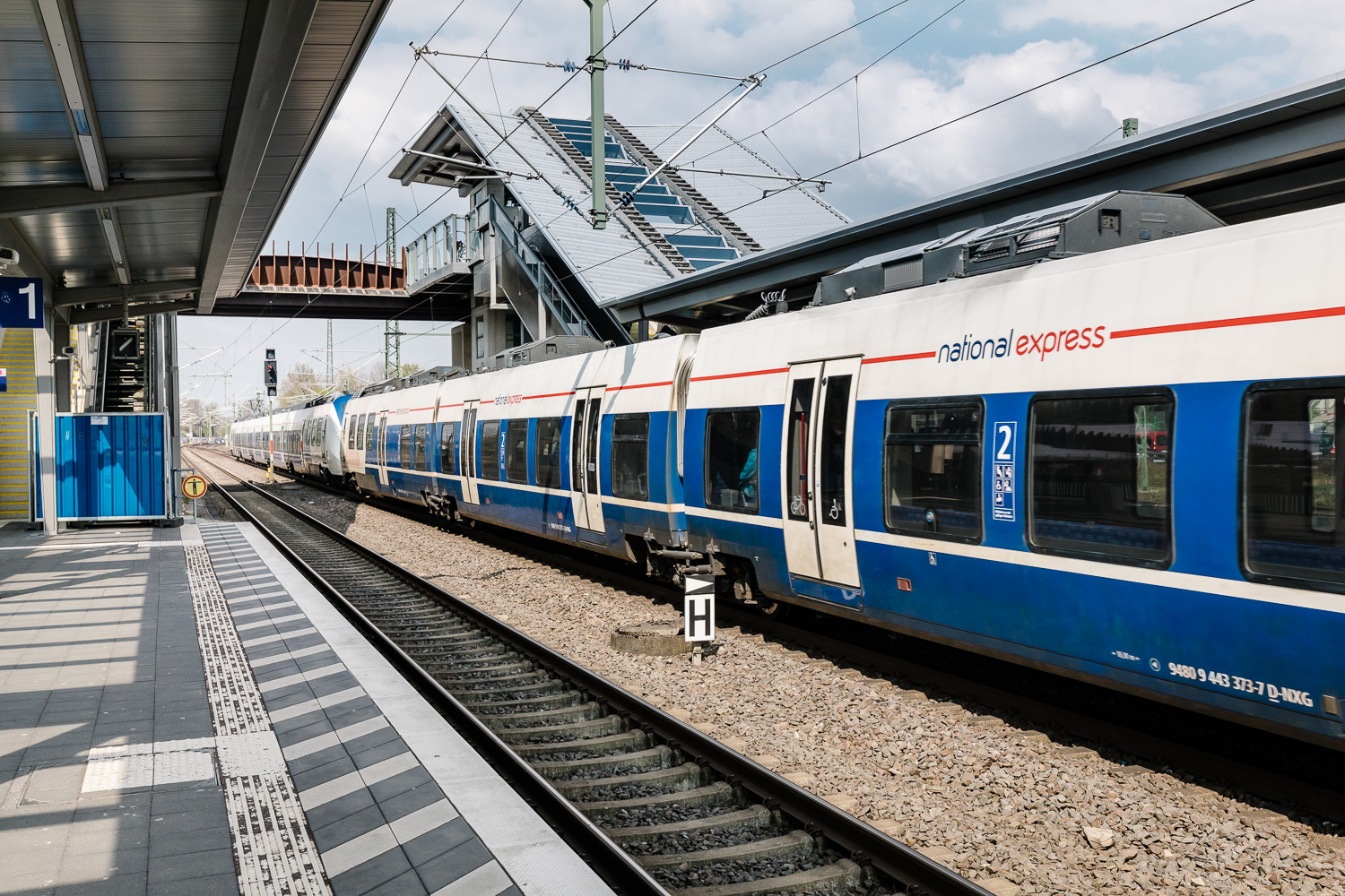 Ein blau-weißer National Express-Zug hält an einem teilweise bewölkten Tag an einem Bahnsteig unter einer Fußgängerbrücke. Der Bahnsteig hat eine Überdachung, Beschilderung und markierte Linien auf dem Gehweg.