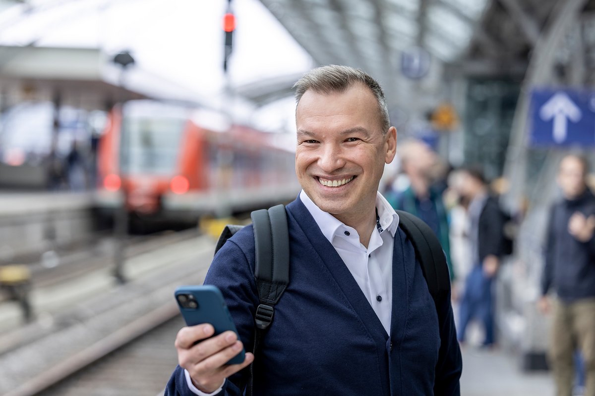 Mann schaut in ein Handy vor einfahrendem Zug im Hintergrund, am Kölner Hauptbahnhof (© go.Rheinland / Smilla Dankert)